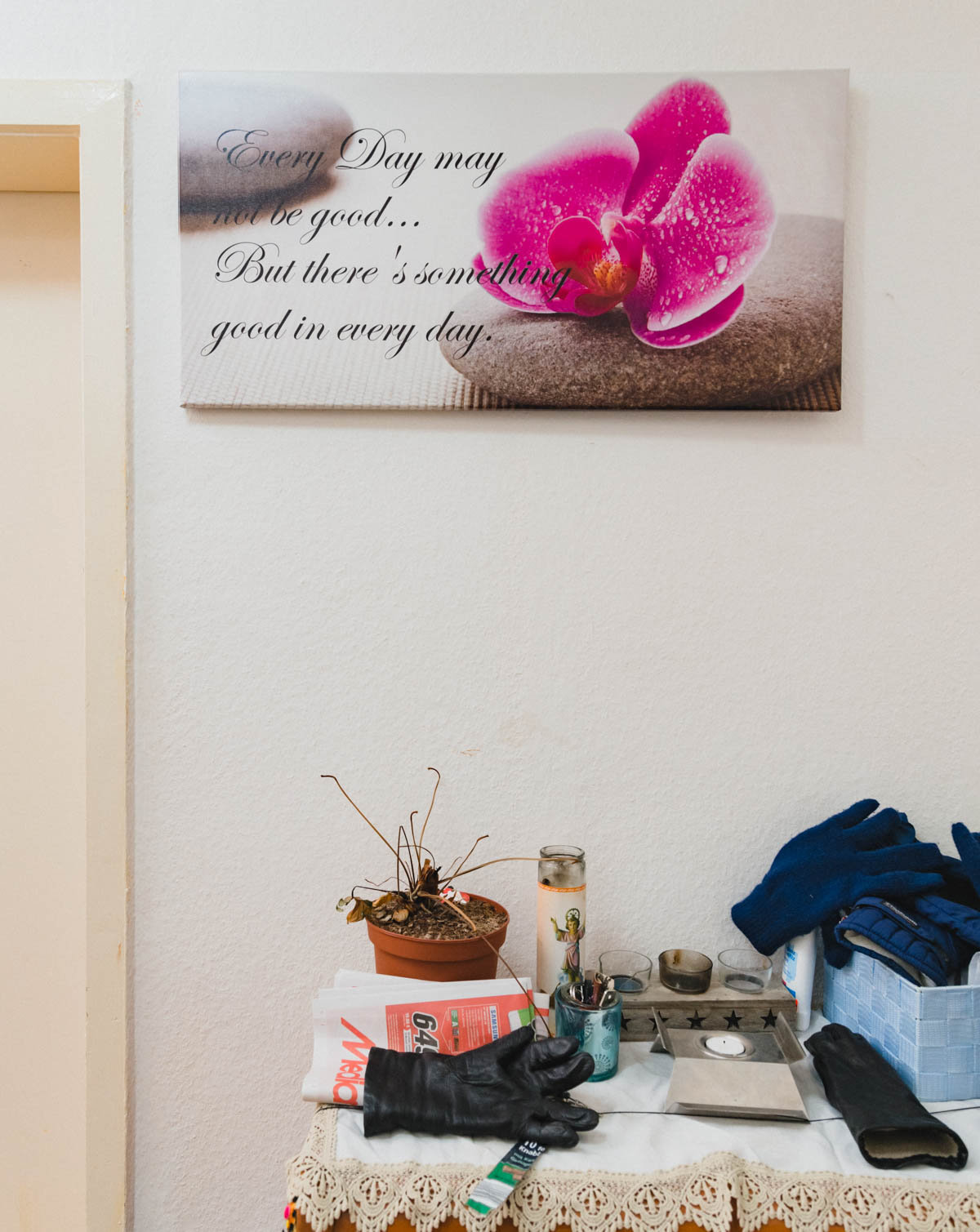 Die Wand einer Wohnung: Ein Bild einer pinken Blüte, verziert mit einer Lebensweisheit, darunter eine Ablage, auf der unter anderem Handschuhe, Teelichter und ein Werbeprospekt liegen   (Foto: Christian Protte)