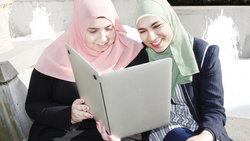 Zwei junge Frauen mit Kopftuch schauen auf einen Laptop, als wäre es ein Buch
