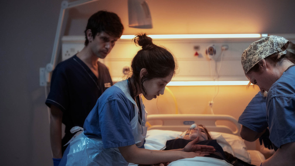 Adam (Ben Whishaw) beobachtet im Hintergrund, wie Shruti (Ambika Mod) eine Patientin in einem Krankenbett versorgt Adam (Ben Whishaw) beobachtet im Hintergrund, wie Shruti (Ambika Mod) eine Patientin in einem Krankenbett versorgt